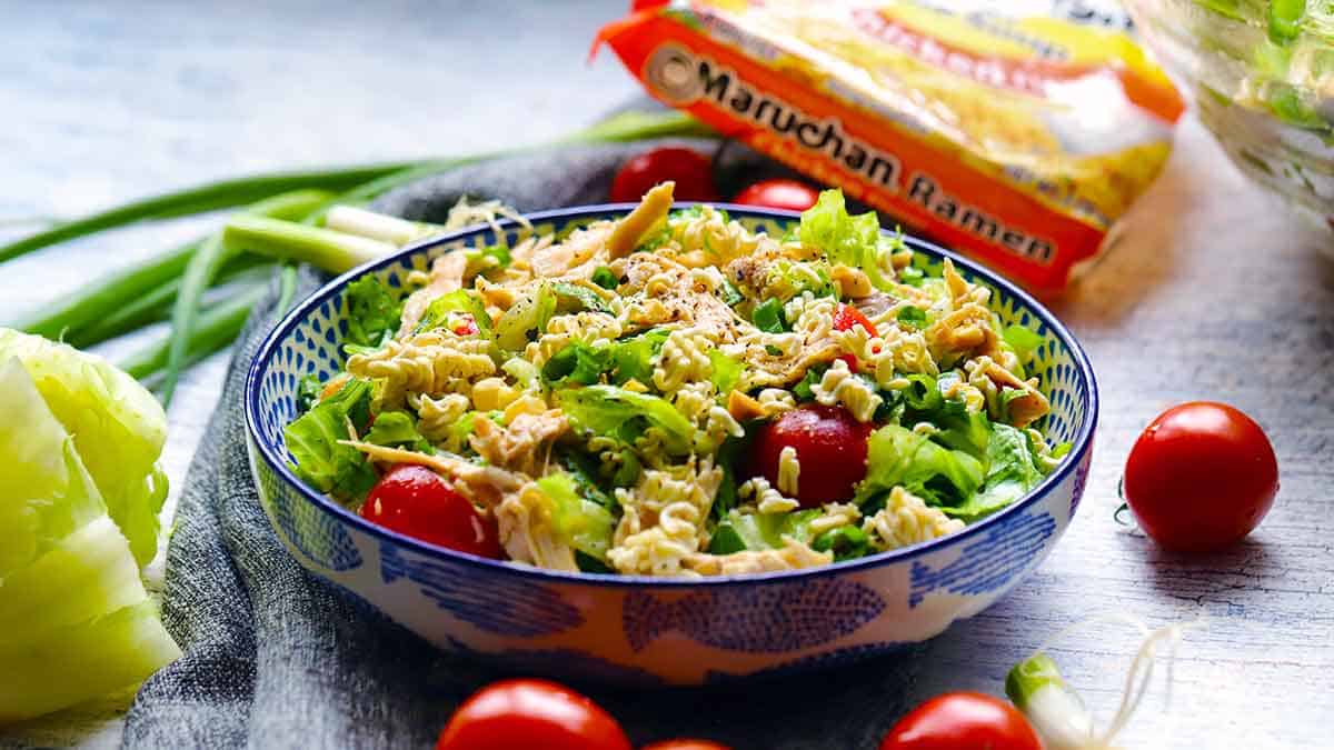 Side view of a bowl of crunchy ramen noodle salad with ingredients and ramen package wrapper scattered around.