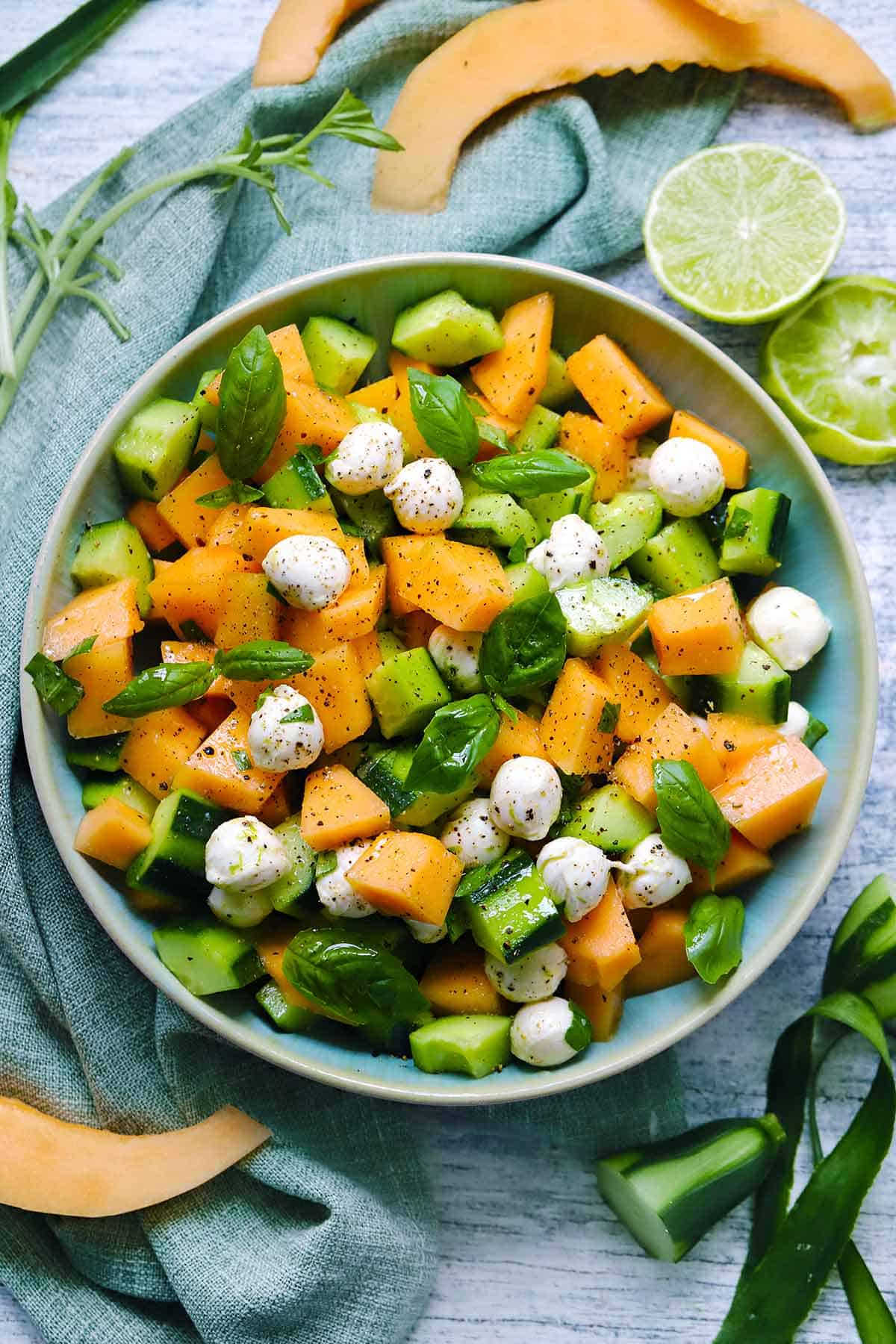Overhead view of a bowl of cucumber melon salad with fresh basil and mozzarella cheese, with scraps of cucumbers, limes, and cantaloupe slices scattered around.