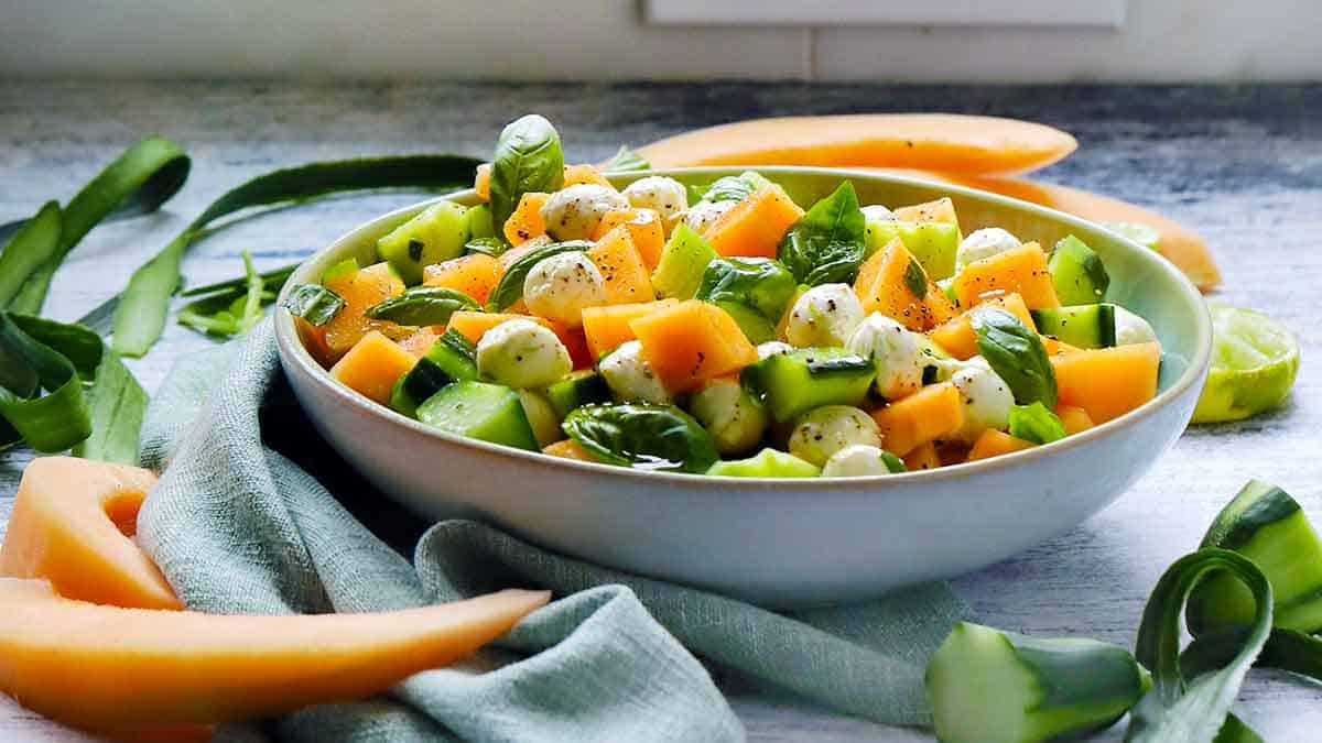 Side view of a bowl of cucumber melon salad, with slices of cantaloupe and scraps of cucumber scattered around.