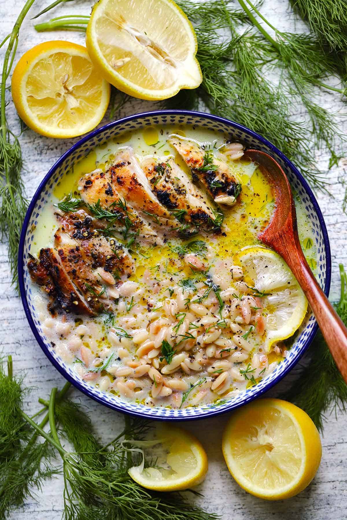 Overhead view of a broth bowl with sliced broiled souvlaki chicken, orzo, and lemony avgolemono broth, garnished with dill, surrounded by lemon halves and fresh dill.