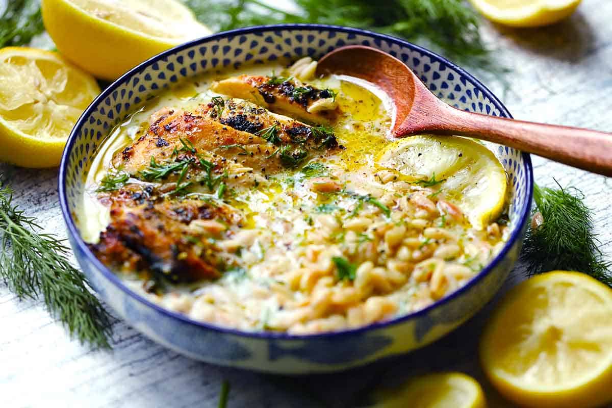 Angled close-up of a bowl of chicken souvlaki avgolemono broth bowl with sliced broiled souvlaki chicken, orzo, and creamy lemon broth, with a wooden spoon and lemon slices.