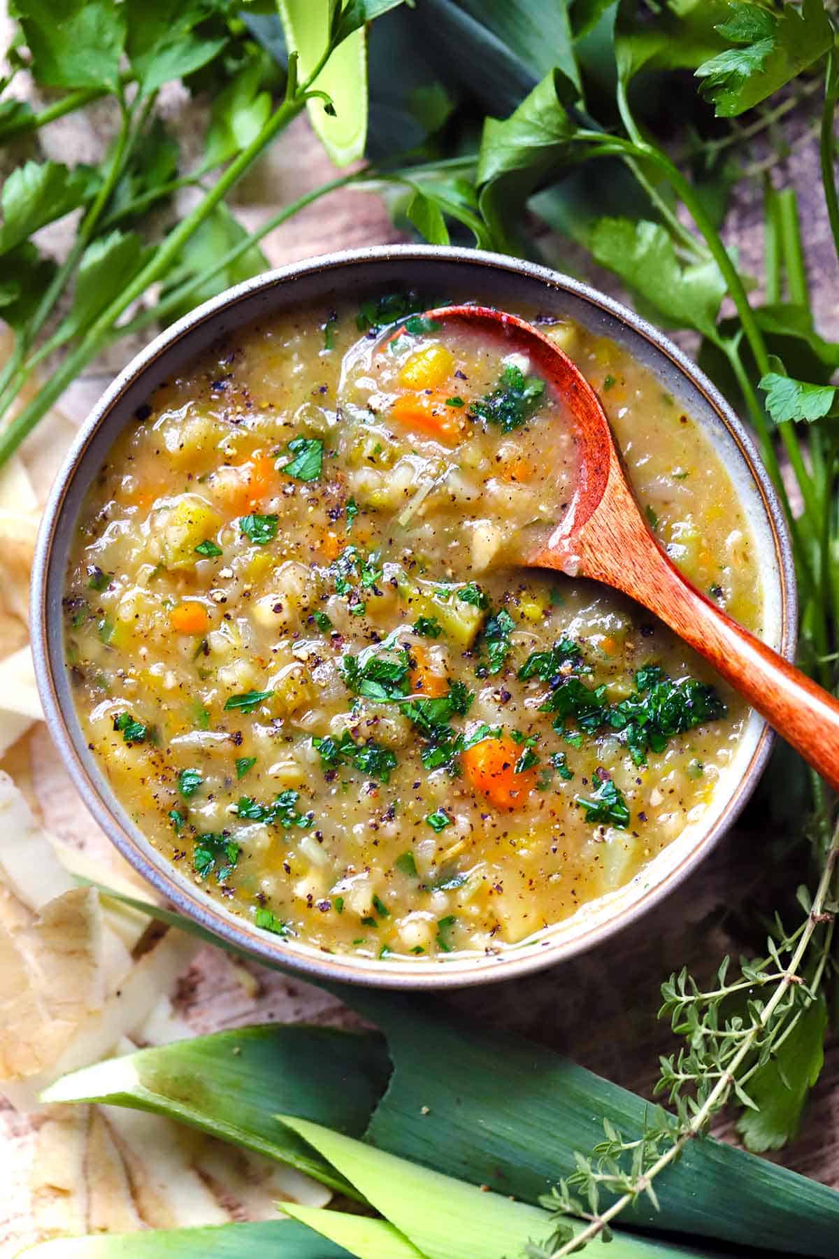 Overhead view of rustic vegetable barley soup with carrots, barley, and chopped parsley in a gray bowl with a wooden spoon, surrounded by leeks, fresh herbs, and thyme.