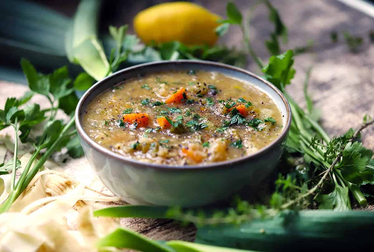 Angled view of a bowl of vegetable barley soup with carrots and parsley, surrounded by leeks, fresh parsley, thyme, and a lemon.
