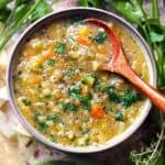 Square overhead photo of vegetable barley soup with carrots, barley, and parsley in a gray bowl with a wooden spoon, surrounded by fresh herbs.